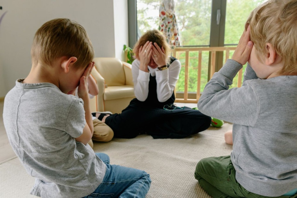 Three children playing peekaboo in a room with large windows, enjoying togetherness indoors.